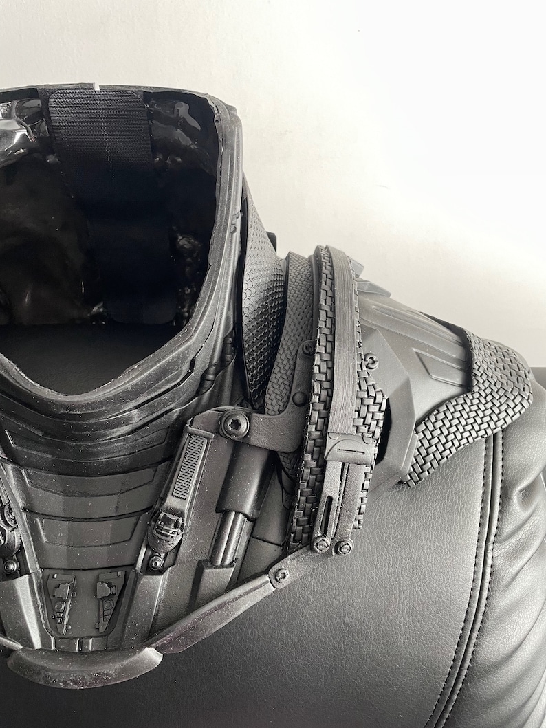 May include: Black and gray plastic costume piece with a textured surface and a series of buckles and straps. The piece appears to be a neck and shoulder armor piece.