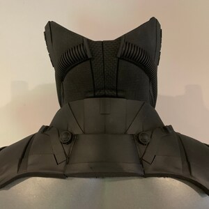 May include: A black, futuristic-looking neck and shoulder armor piece. The armor has a textured surface and is designed with multiple panels and rivets. The design includes a high collar and shoulder guards, suggesting a protective function.