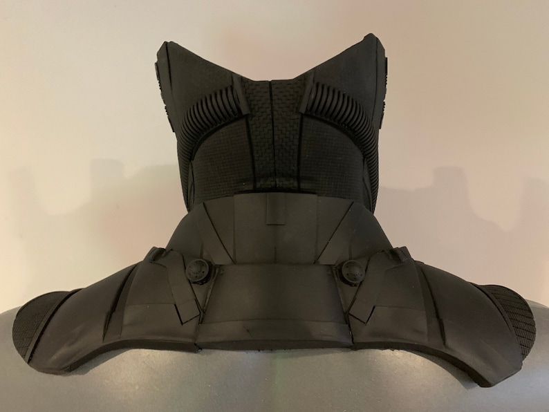 May include: A black, futuristic-looking neck and shoulder armor piece. The armor has a textured surface and is designed with multiple panels and rivets. The design includes a high collar and shoulder guards, suggesting a protective function.
