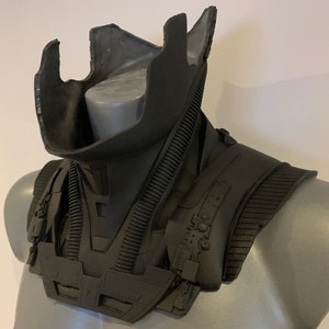 May include: A black, futuristic-looking neck and shoulder armor piece. The armor has a layered design with ribbed and textured sections. It is displayed on a gray mannequin, showcasing its shape and details.