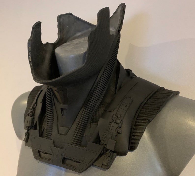 May include: A black, futuristic-looking neck and shoulder armor piece. The armor has a layered design with ribbed and textured sections. It is displayed on a gray mannequin, showcasing its shape and details.