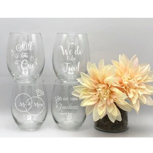 May include: Four clear glass wine glasses with etched designs. The glasses are arranged in a row with flowers in a vase behind them. The designs on the glasses include "Still the One", "We do take two!" , "Mr. & Mrs." and "Let's eat, drink & get remarried".
