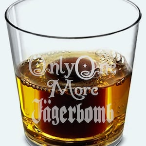 May include: A clear glass tumbler filled with a dark amber liquid, possibly a cocktail. The glass has the words "Only One More J&auml;gerbomb" etched in a decorative font. The drink is partially filled with a darker, reddish-brown liquid.