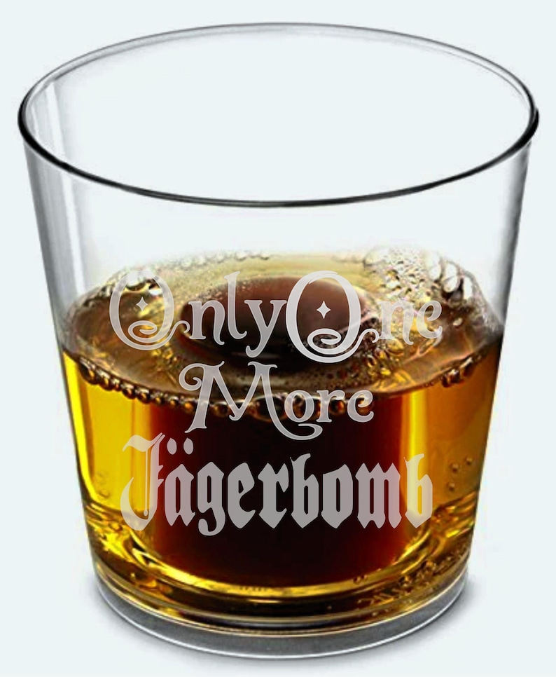 May include: A clear glass tumbler filled with a dark amber liquid, possibly a cocktail. The glass has the words "Only One More J&auml;gerbomb" etched in a decorative font. The drink is partially filled with a darker, reddish-brown liquid.