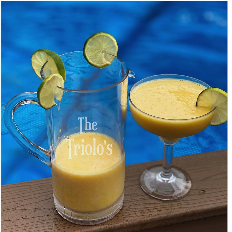 Puede incluir: Una jarra de vidrio transparente con asa, llena de un l&iacute;quido amarillo y decorada con el texto "The Triolo's". Un vaso de margarita lleno de un l&iacute;quido amarillo est&aacute; al lado de la jarra. Ambas bebidas est&aacute;n adornadas con rodajas de lima.