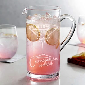 May include: A clear glass pitcher with a handle filled with a pink drink and ice. The pitcher has the words "Cosmopolitan cocktail" and the ingredients "Cointreau, cranberry juice, lime juice, vodka" etched on the side.