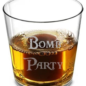May include: A clear glass tumbler with the words "Bomb Party" etched on the side. The glass is filled with a dark amber liquid, possibly a cocktail or spirit, with a foamy top.