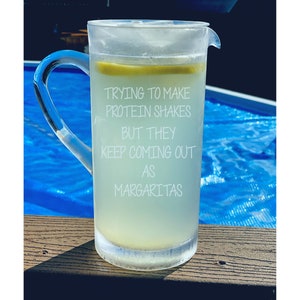 May include: A glass pitcher filled with a light yellow liquid and a lemon slice. The pitcher has a white inscription that reads "Trying to make protein shakes but they keep coming out as margaritas."
