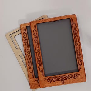 Puede incluir: Tres marcos de fotos de madera. Dos están teñidos de color marrón rojizo y uno es de color madera clara natural. Los marcos tienen forma rectangular con diseños florales decorativos grabados. El centro de los marcos es de color gris oscuro.