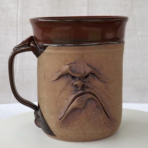 May include: A handmade ceramic mug with a sculpted, grumpy face design. The mug is primarily tan with a dark brown rim and handle. The handle has a unique, textured design. A unique, handcrafted piece for coffee or tea.