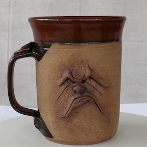 May include: A handmade ceramic mug with a sculpted, angry face design. The mug features a dark brown rim and handle, contrasting with the tan body. The face is deeply carved, adding texture and character to this unique piece. A functional and artistic item.