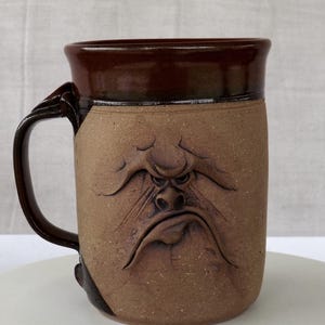 May include: A stoneware mug with a dark brown rim and handle. The mug features a carved, grumpy face design on a tan background. The handle has a unique, textured design.