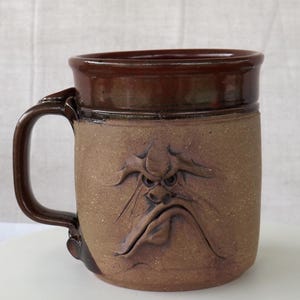 May include: A brown ceramic mug with a handle, featuring a carved, grumpy face. The mug has a dark brown rim and handle, with a lighter brown body. The face has furrowed brows and a downturned mouth.