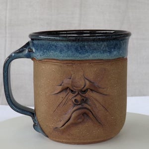 May include: A handmade ceramic mug with a grumpy face design. The mug features a brown body with a blue rim and handle. The face is sculpted with furrowed brows, a downturned mouth, and a textured surface, creating a unique and expressive piece.