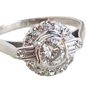 May include: A silver ring with a large round diamond surrounded by smaller diamonds. The ring has a delicate band and a halo setting.