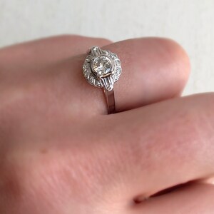 May include: A silver ring with a round, clear diamond center stone surrounded by smaller diamonds. The ring has a delicate, vintage-inspired design.