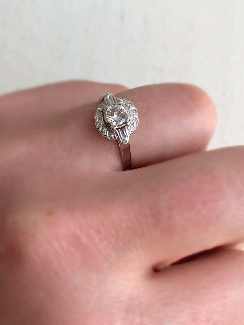 May include: A silver ring with a round, clear diamond center stone surrounded by smaller diamonds. The ring has a delicate, vintage-inspired design.