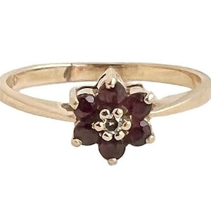May include: A gold-toned ring featuring a flower-shaped design. The flower is composed of dark red gemstones, with a smaller, lighter-colored center. The ring's band is a simple, polished gold.