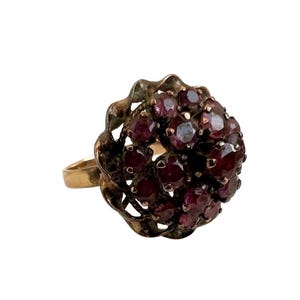 May include: A gold-toned ring featuring a cluster of small, deep red gemstones. The gemstones are set in a floral-shaped mounting with an openwork design. The ring has a classic, vintage aesthetic.