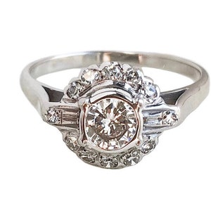 May include: A silver ring with a large round diamond surrounded by smaller diamonds. The ring has a delicate design with a halo of smaller diamonds around the center stone.