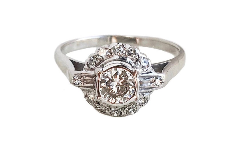 May include: A silver ring with a large round diamond surrounded by smaller diamonds. The ring has a delicate design with a halo of smaller diamonds around the center stone.
