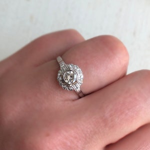 May include: A silver ring with a round, clear diamond center stone surrounded by a halo of smaller diamonds. The band is set with three baguette diamonds.