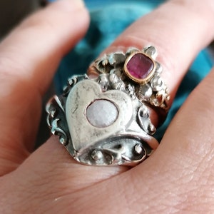 May include: A silver ring with a heart-shaped design featuring a white stone. The ring is worn on a finger with a second ring featuring a pink stone.