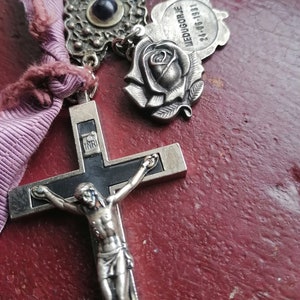 Op de afbeelding: Een zilveren en zwart kruisbeeld met een figuur van Jezus Christus op het kruis. Het kruisbeeld is bevestigd aan een ketting met verschillende bedels, waaronder een roos, een klein oogbedeltje en een bedeltje met de tekst "MEDUGORJE 1981-06-24".