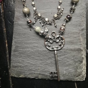 Puede incluir: Un collar plateado con un colgante de llave, un colgante de corazón con cerradura y varias cuentas. El collar está formado por una cadena y una hebra de cuentas.