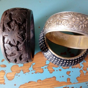 Puede incluir: Tres pulseras decorativas, una de madera negra con diseño floral tallado, una de plata con diseño floral en relieve y una de color negro con un patrón geométrico.