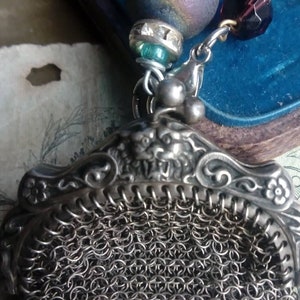 May include: A silver chainmail purse with a decorative silver clasp. The purse is hanging from a chain with various beads, including a purple and iridescent bead, a clear bead with rhinestones, and a dark red bead.