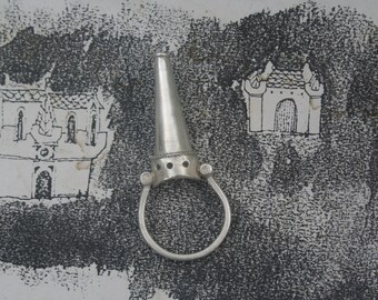 May include: A silver ring with a pointed cone-shaped top. The ring has a circular band and a decorative design around the base of the cone.