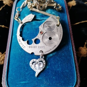 May include: A silver necklace with a heart-shaped pendant featuring a clear rhinestone. The pendant is attached to a circular piece of metal with the words "HEART & COLD", "LAKE SHORE", and "21 JEWELS" engraved on it.