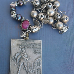 以下が含まれることがあります： 銀色のネックレスに、歴史的な衣装を着た男性の銀色のレリーフが描かれたペンダントが付いています。ペンダントには、「VIAREGGIO 10 FEBBRAIO 1900」と「Lo Straviareggio Club tende omaggio alla maschera genovese di Capitan Spaventa e a tutta la Ligur a Genova Porta Toprana e la Casa Colombo」というテキストが書かれています。