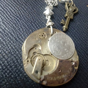 Può includere: Una collana con catena d'argento con un ciondolo a forma di quadrante dell'orologio in ottone, un ciondolo a forma di chiave d'argento e un ciondolo a forma di fiore d'argento. Il quadrante dell'orologio ha una moneta d'argento sopra.