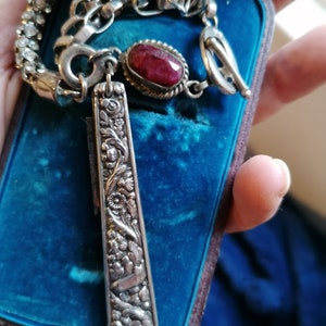 May include: A silver necklace featuring a red gemstone pendant and a detailed silver charm. The charm is shaped like a long, thin blade with an intricate floral design.