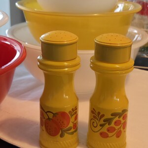 May include: Two yellow salt and pepper shakers with fruit designs, alongside a collection of mixing bowls in red, white, and yellow. The shakers have a textured base and a perforated top. The bowls are stacked, suggesting kitchen storage.