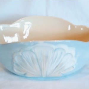 May include: A light blue ceramic bowl with a scalloped rim and a white seashell design on the exterior. The interior is a light beige color. This decorative bowl is suitable for serving or display.
