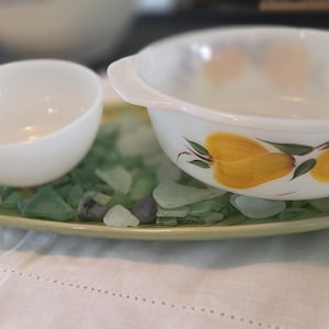 May include: A white bowl and a small white dish sit on a green and yellow tray filled with sea glass. A larger white bowl with yellow floral designs is also on the tray. The items are on a white tablecloth.
