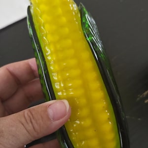 May include: A decorative glass corn on the cob sculpture. The corn is a bright yellow colour, encased in green glass husks. The sculpture is held by a hand.