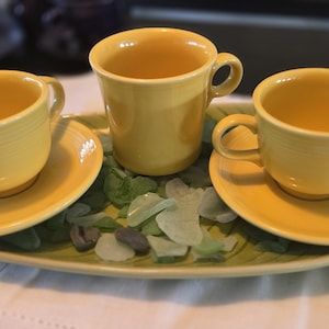May include: Three yellow teacups and saucers are arranged on a green, leaf-shaped tray, with decorative sea glass. The cups have handles and sit on matching saucers. The set is a cheerful, bright yellow.