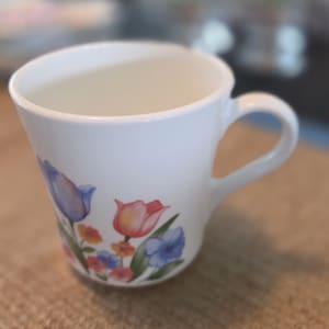 May include: White ceramic coffee mug with a curved handle. The mug is decorated with a floral design featuring blue, pink, and orange tulips and other flowers. The mug is sitting on a brown woven surface.