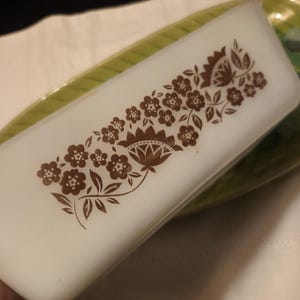 May include: A rectangular, white glass dish with a brown floral pattern. The design features stylized flowers and leaves. The dish is resting on a green and white plate, suggesting a serving or decorative purpose.