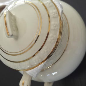 May include: Ivory-colored porcelain teapot with gold trim. The lid and body feature concentric gold bands and embossed detailing. The handle and spout are also accented with gold. A white cloth is wrapped around the spout.