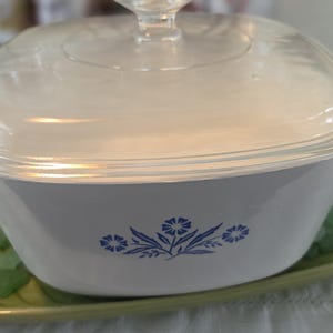 May include: White casserole dish with a clear glass lid, featuring a blue floral design. The dish is set on a green, leaf-shaped plate with decorative sea glass. A classic kitchenware item.