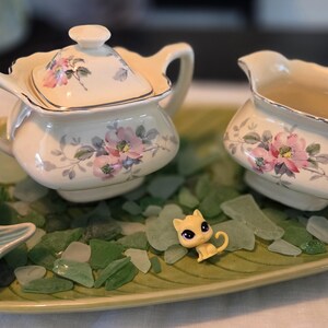 May include: A cream-colored tea set with floral designs, including a teapot and creamer, sits on a leaf-shaped tray. A small pink rubber duck and a yellow cat figurine are also on the tray, surrounded by green glass pieces.