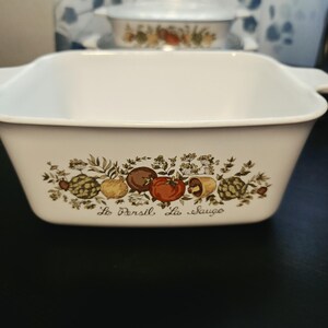May include: A white square ceramic baking dish with integrated side handles. The front features a colorful botanical illustration of vegetables and herbs, including tomatoes and mushrooms, with the text Le Persil La Sauge printed below.