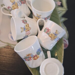 May include: Set of six white ceramic teacups with handles. Each cup features a colorful illustration of a teddy bear holding a gift box. The cups are arranged on a green and white leaf-shaped plate.