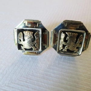 Può includere: Un paio di orecchini in argento e oro con un design geometrico che presenta una figura di animale stilizzata. Gli orecchini sono di forma quadrata con un bordo dorato e uno sfondo nero.