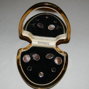 May include: A gold-colored box with a black velvet interior, containing a set of eight pearl cufflinks. The box is open and the cufflinks are arranged in the velvet lining. The box has the word "Krementz" printed on the inside.